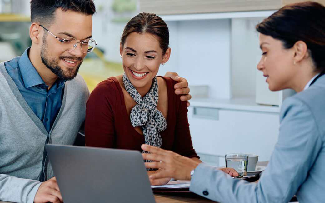 happy couple using laptop with car saleswoman in showroom